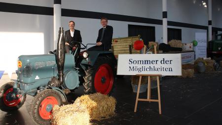 Ministerpräsident Daniel Günther und Christian Kuhnt sitzen auf einem Traktor in einer großen Halle. Um sie herum liegen einige Strohballen herum, auf einem Schild steht 
